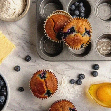 Golden-brown homemade muffins recipe with fresh blueberries in muffin tin surrounded by baking ingredients