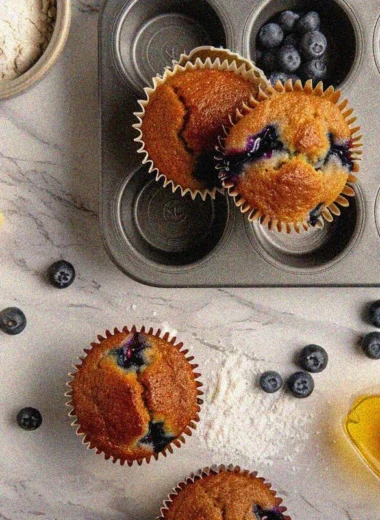 Golden-brown homemade muffins recipe with fresh blueberries in muffin tin surrounded by baking ingredients